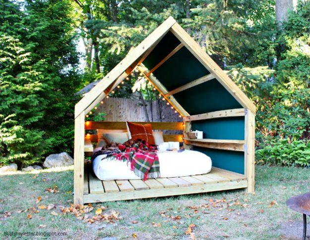 Outdoor cabana with a gabled open frame painted dark inside, string lights and a daybed on a slatted deck floor