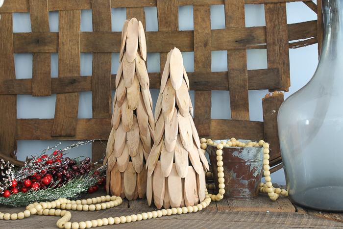 Two cone Christmas trees covered in overlapping pale wood slices beside a wood bead garland, red berries and a rusty bucket