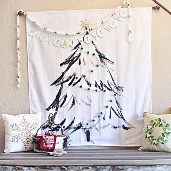 A white fabric wall hanging painted with a black brush stroke tree and green pom pom ornaments hung from a rod above a bench