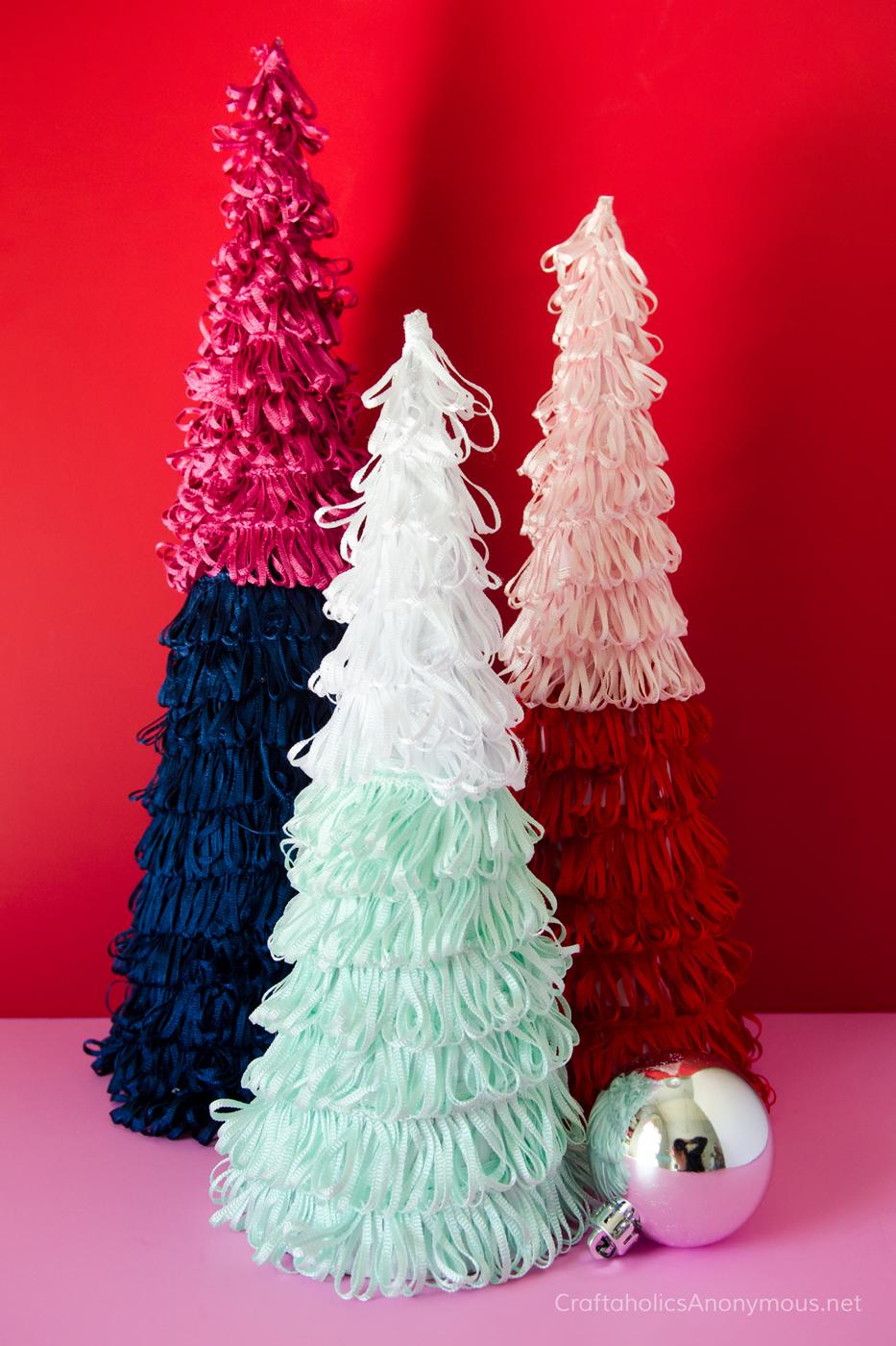 Three cone Christmas trees covered in loopy ruffle trim in pink and navy, white and mint and blush and red against a red wall