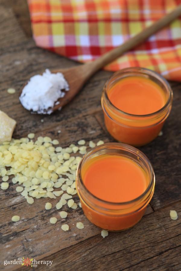 Turmeric pain relief balm, two small jars of orange balm on weathered wood with beeswax pellets and a wooden spoon