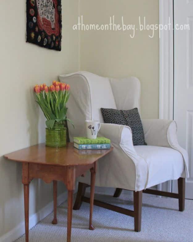 Wing chair covered in a creamy white drop cloth slipcover with piped edges, beside a small round wooden table