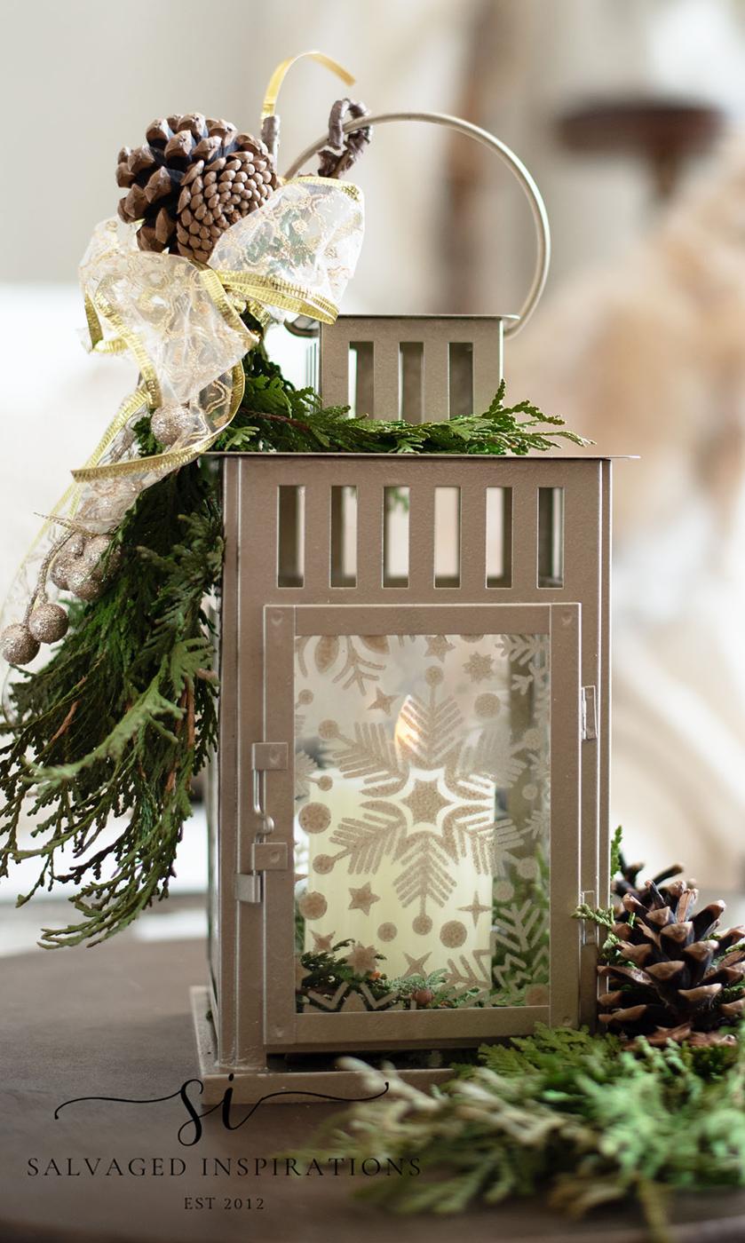 A champagne painted lantern with stenciled snowflake glass, a pillar candle inside, greenery, pine cones and gold ribbon