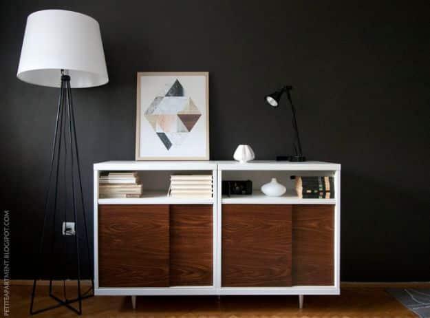 Long white cabinet with brown faux wood sliding doors on slim metal legs against a charcoal wall, lamp and geometric print