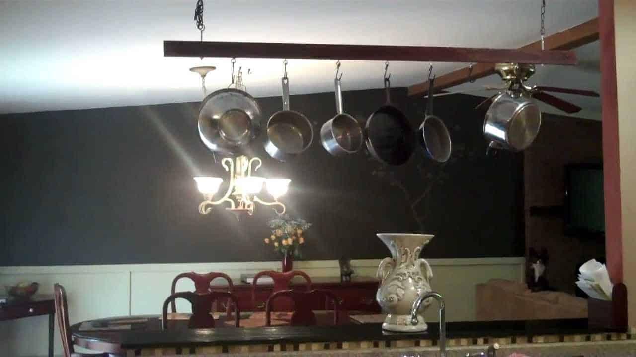 Wooden ceiling pot rack hung on chains over a dining area, holding six stainless pans above a chandelier and dark wall