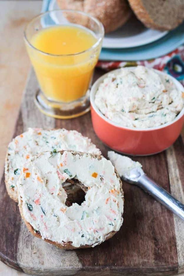 Veggie cream cheese spread on a bagel beside a bowl of the flecked spread and a glass of orange juice