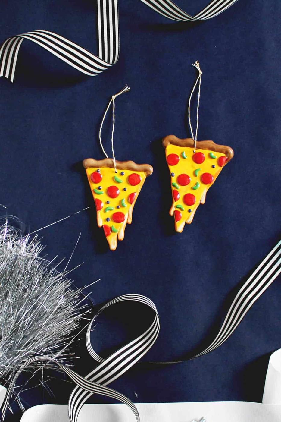 Two painted clay pizza slice ornaments with pepperoni and green peppers hanging on twine loops against navy paper