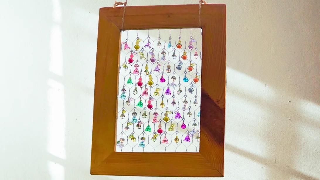 Picture frame suncatcher, a wooden frame strung with rows of tiny colored beads hanging on a pale wall