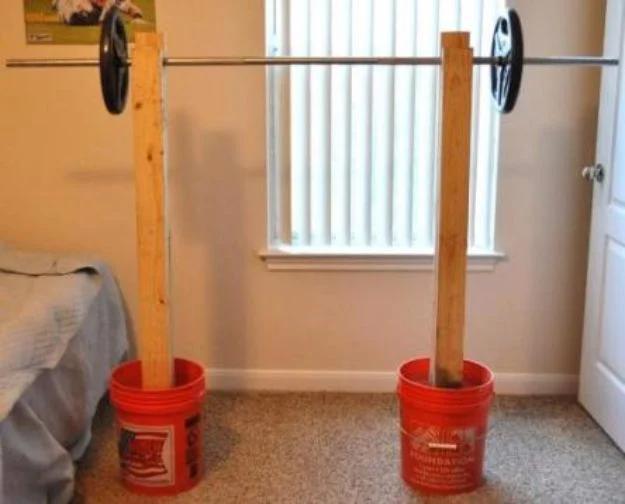 Two homemade squat rack uprights made of stacked two by fours set in red five gallon buckets, holding a loaded barbell