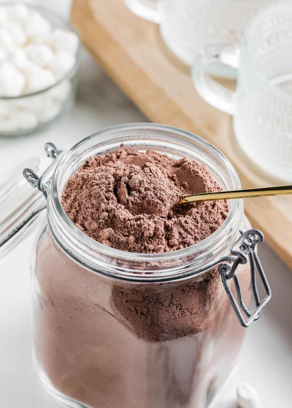 Large clip top jar filled with homemade cocoa mix with a gold spoon in it, marshmallows and mugs behind