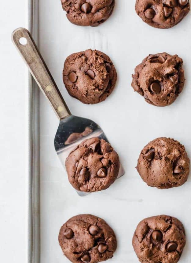 Double chocolate chip cookies cooling on a metal baking sheet with a spatula lifting one from the middle