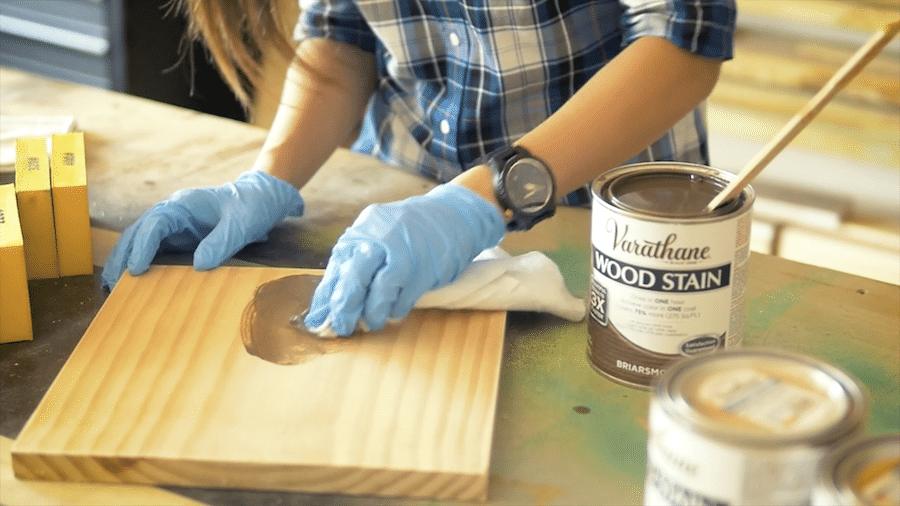 Gloved hands wiping wood stain into a pine board with a rag, stain can and brushes on the workbench behind