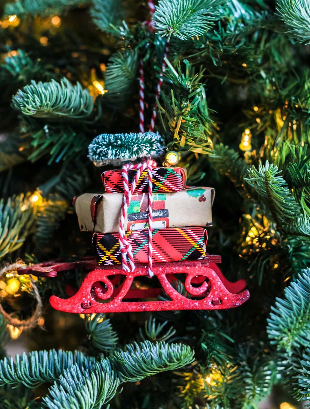 Red glitter sleigh ornament stacked with tiny tartan and kraft wrapped parcels and a bottle brush tree