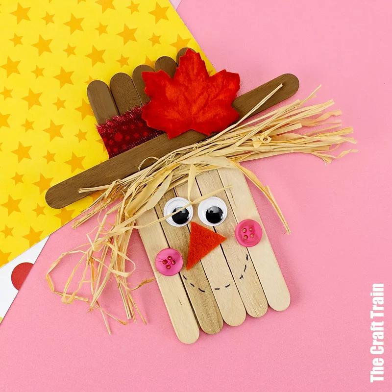 A craft stick scarecrow with googly eyes, an orange felt nose, pink button cheeks, raffia hair and a red maple leaf