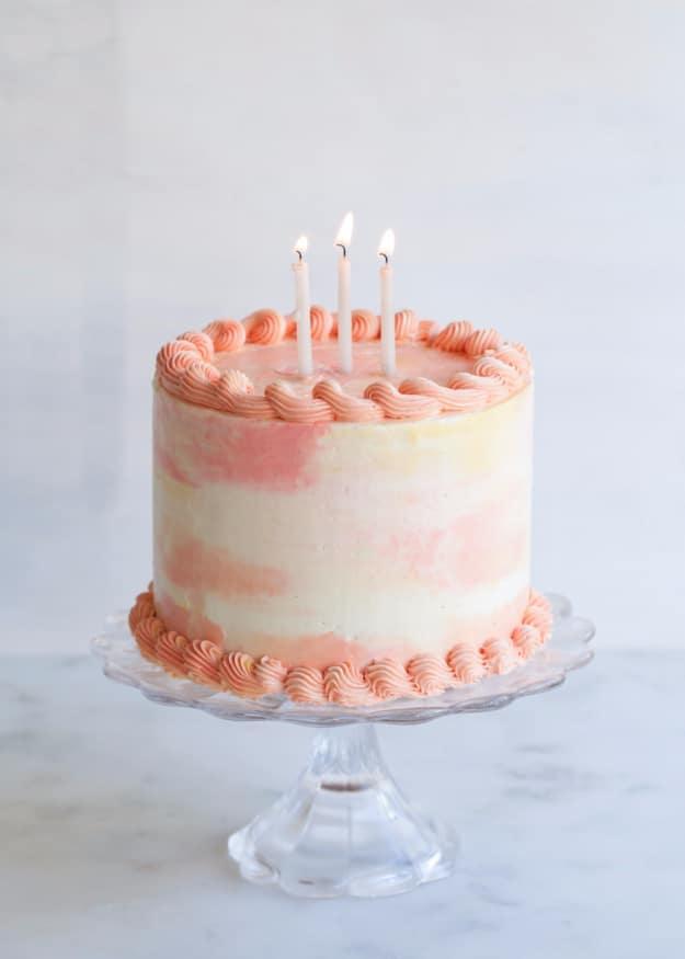 Pale pink and peach watercolor buttercream birthday layer cake with three candles on a glass cake stand