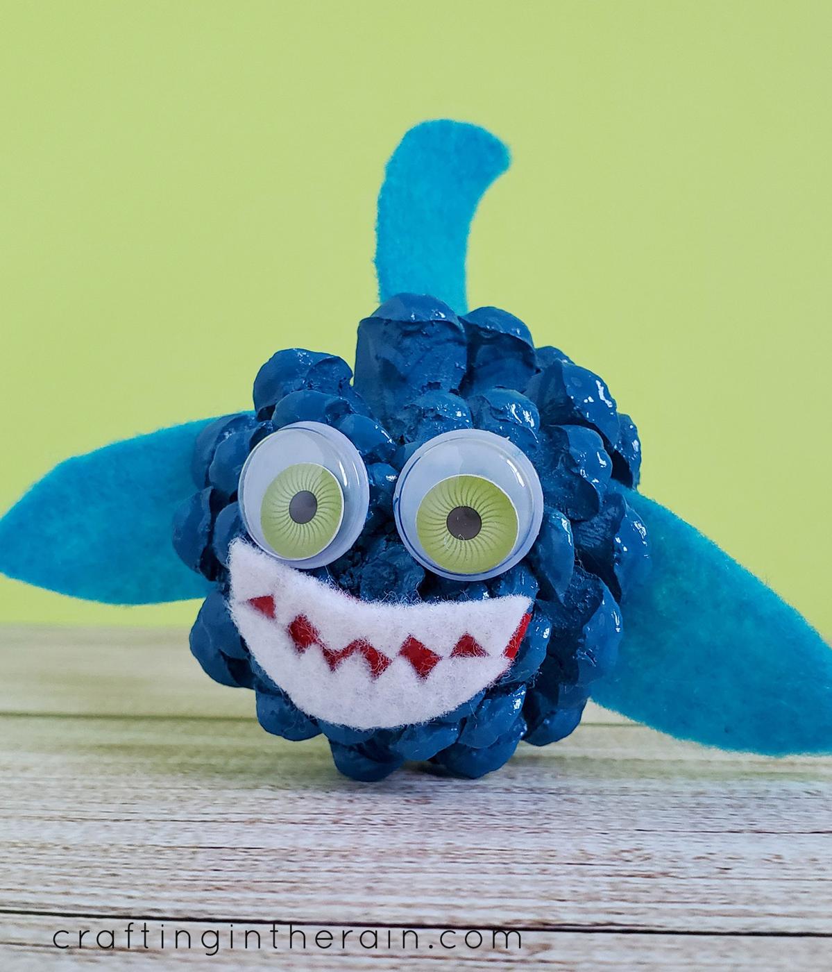 Blue spray painted pine cone shark with felt fins, red mouth, white felt teeth and large googly eyes