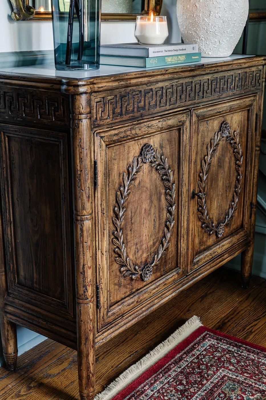 Ornate carved wood cabinet in a warm brown gel stain finish, styled with books, a lantern and a glass vase