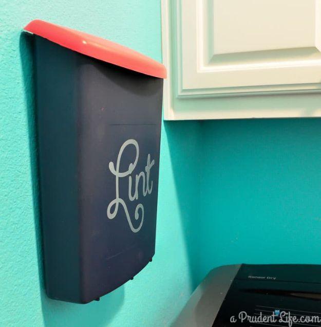 Navy wall mounted lint bin with a coral lid and silver Lint script, hung on a teal laundry wall beside white cabinets