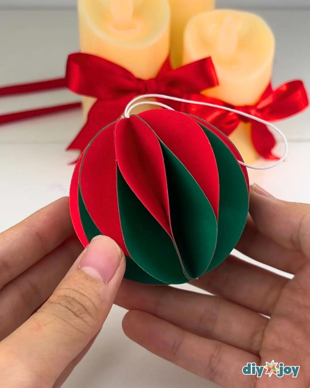 Paper Ornaments - two hands holding a finished red and dark green striped paper ball with a white string loop attached at the top