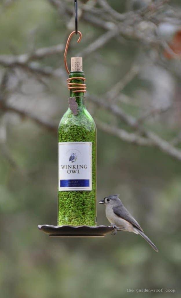 Wine bottle bird feeder hanging from a hook with green seed inside and a small gray bird on the saucer