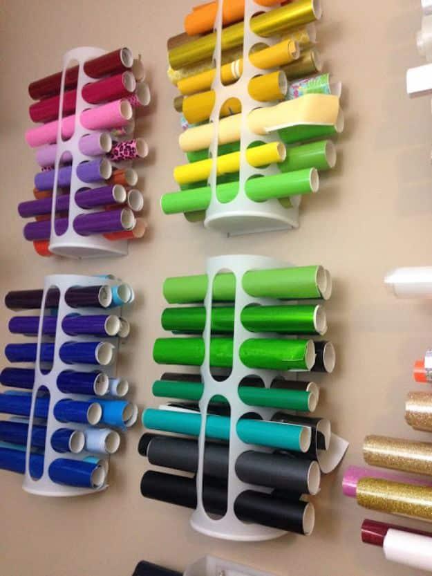 Four white plastic bag holders wall mounted and filled with rolls of craft vinyl sorted by color, more rolls to the side