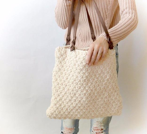 Cream chunky knit tote bag with brown leather handles carried against a pink sweater and pale ripped jeans
