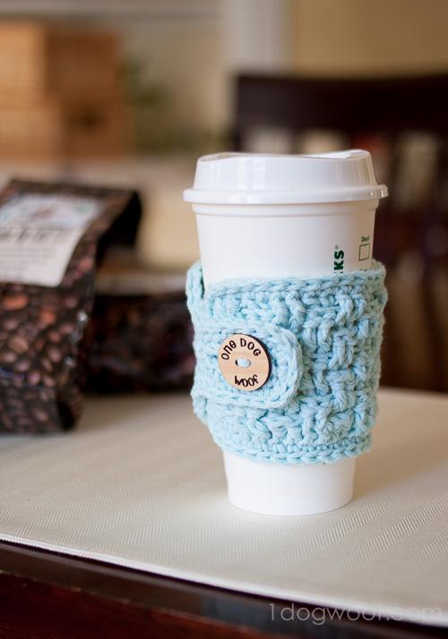 Pale blue crochet basketweave cup cozy buttoned around a white takeout coffee cup on a dark wood table