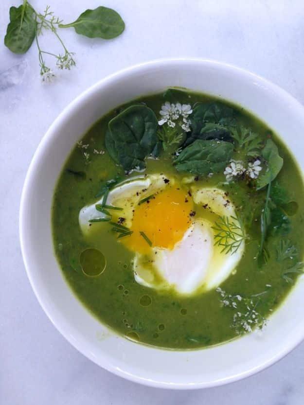 Broccoli spinach soup in a white bowl, bright green with a poached egg, fresh herbs and cracked pepper