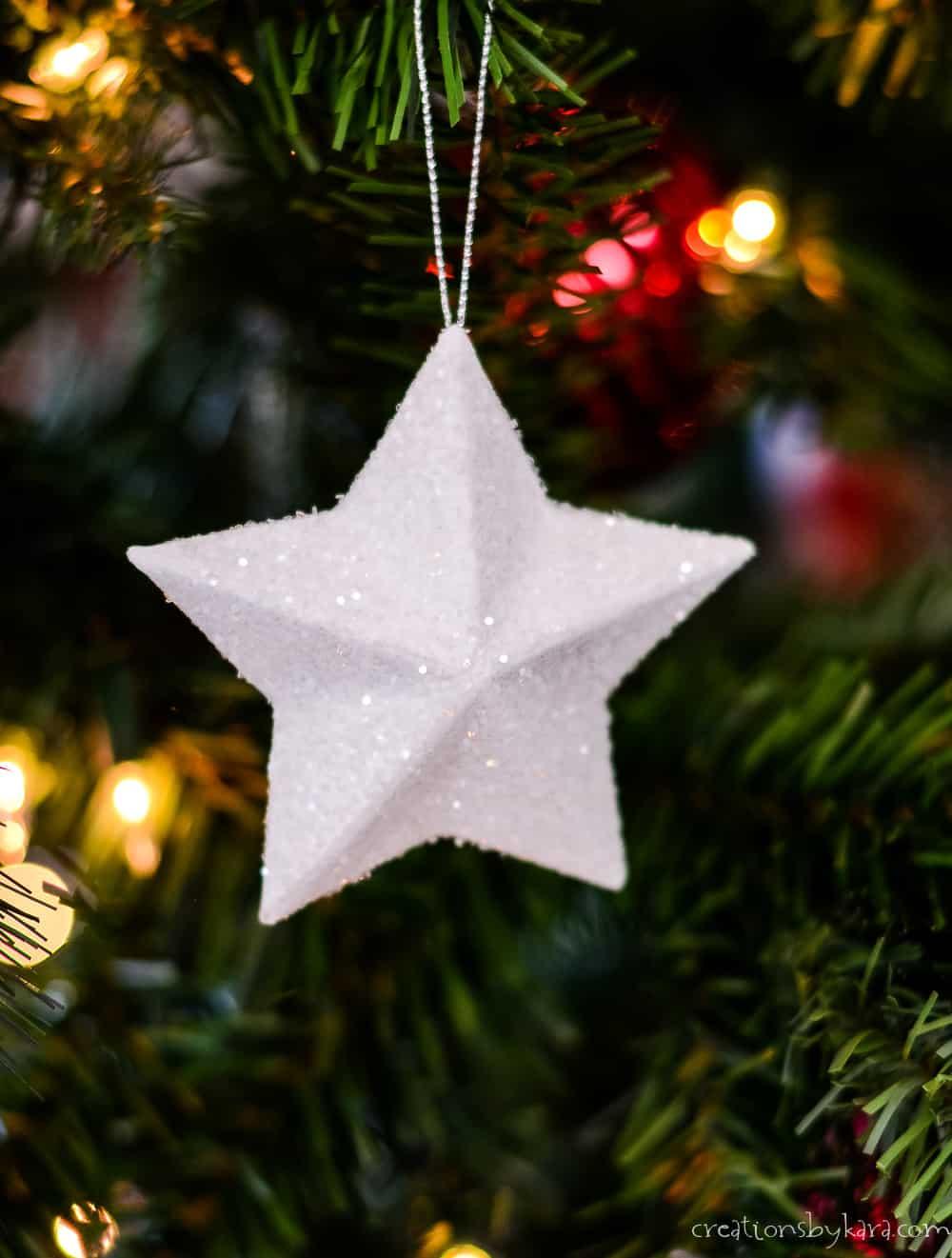 A white glitter three dimensional star ornament hanging on a silver cord in a lit Christmas tree with red and gold bokeh