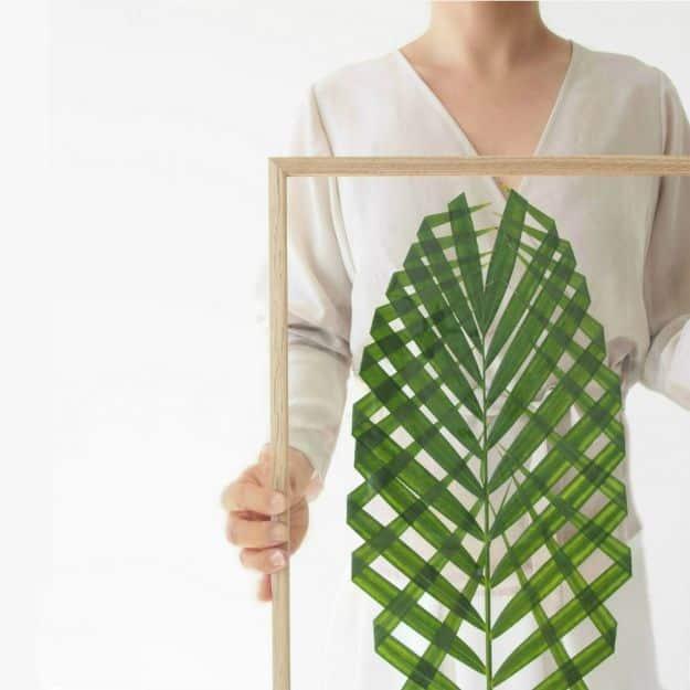 Large woven green paper leaf mounted inside a thin wooden frame, held up by hand against a white wall