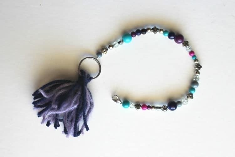 Beaded clip chain in teal, purple and black beads with a purple yarn tassel and a key ring on a white surface