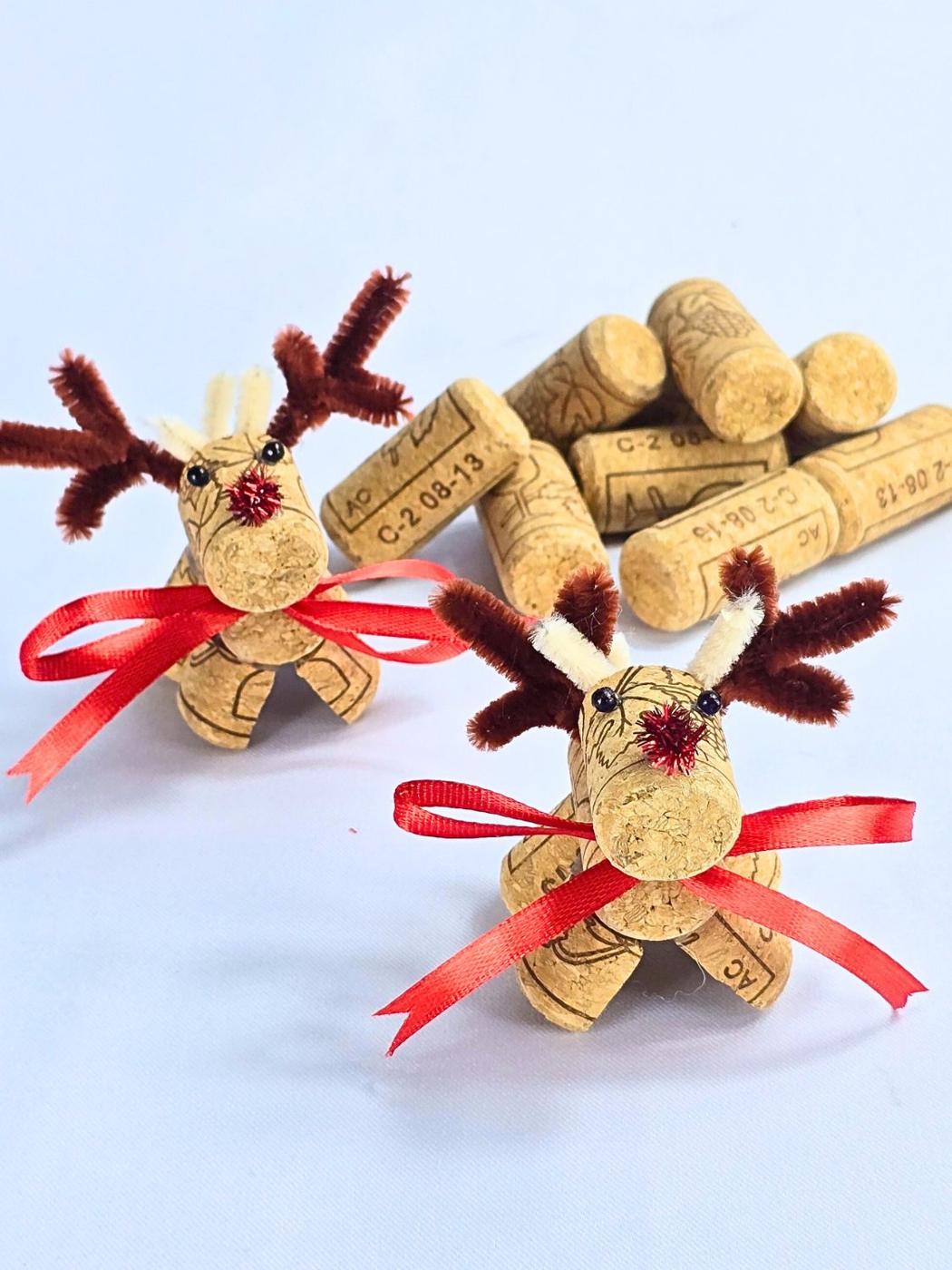 Two wine cork reindeer with brown pipe cleaner antlers, red pom pom noses and red ribbon scarves beside a pile of corks