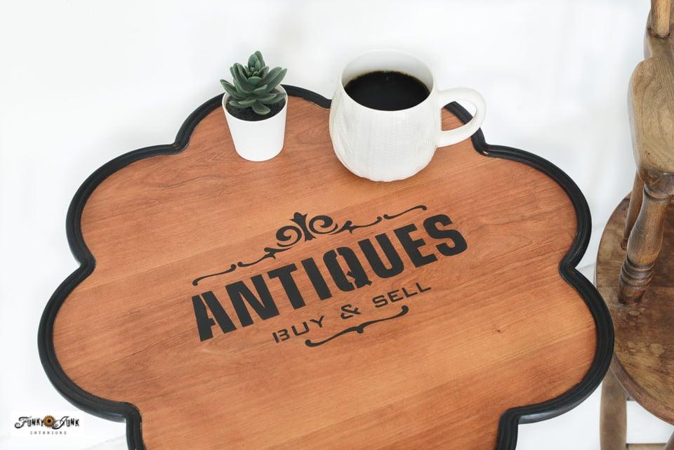 Scalloped pedestal side table with a stained wood top stenciled with an Antiques Buy and Sell sign, styled with a mug