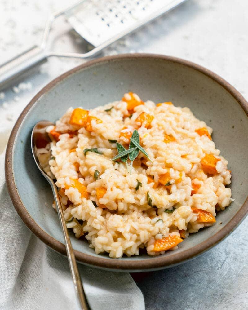 Butternut squash risotto in a grey stone bowl with orange squash cubes, a fresh sage leaf and a spoon resting in it