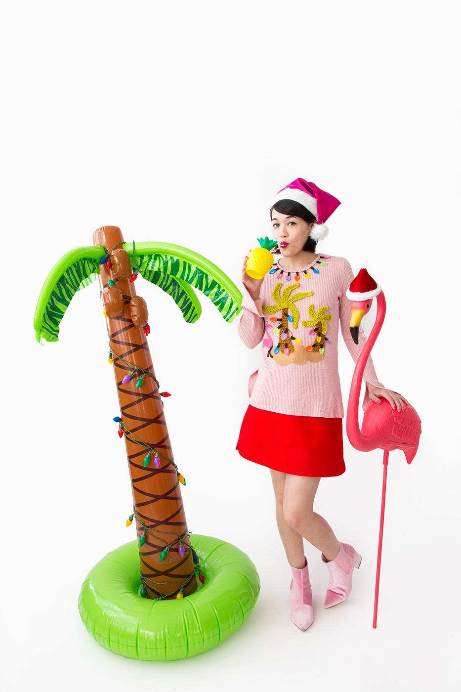 A woman in a santa hat wearing a pink sweater with crocheted palm trees, felt string lights, a flamingo and felt sand