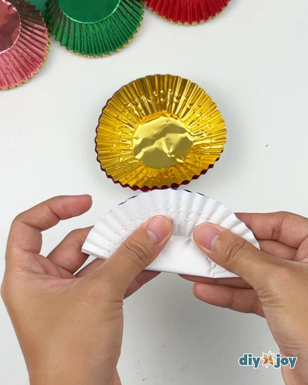 How to Make Christmas Ornaments - a hand folding a white cupcake liner in half beside an upside down gold foil liner