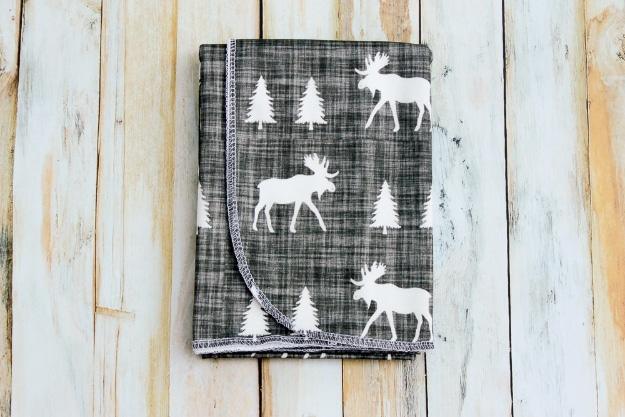Folded charcoal knit swaddle blanket printed with white moose and pine trees, on whitewashed wood boards