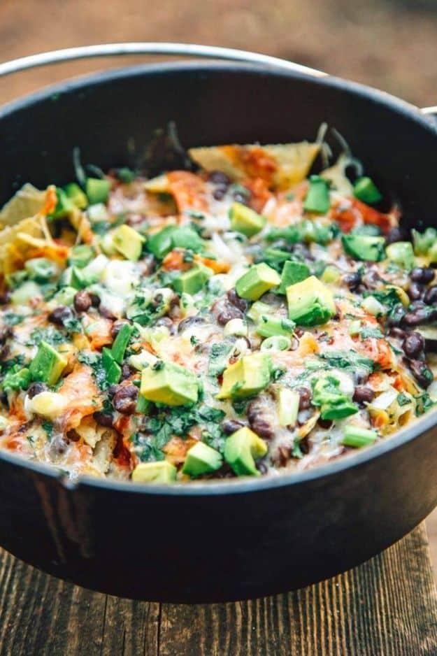 Campfire nachos layered in a dutch oven with black beans, cheese, avocado and cilantro over the fire