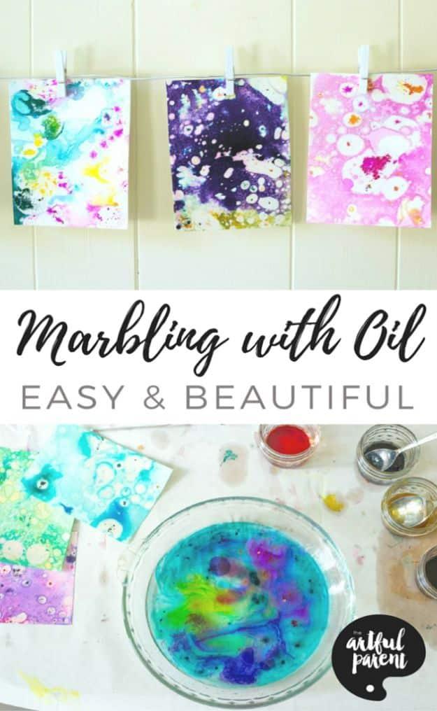 Three marbled paper prints in purple, blue and pink pegged on a line above a bowl of swirled oil and food coloring