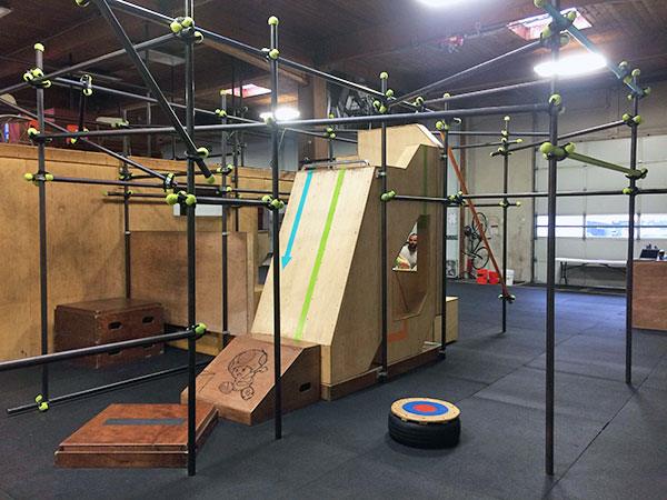 Indoor parkour gym of black steel pipe frames with plywood vault ramps, platforms and a target, on rubber matting