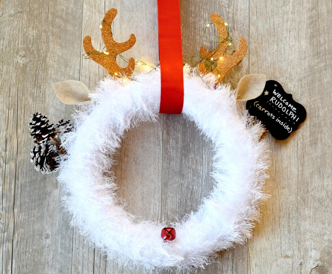 A fluffy white yarn wreath with glitter gold antlers wound with fairy lights, felt ears, a red bell nose and a chalkboard tag