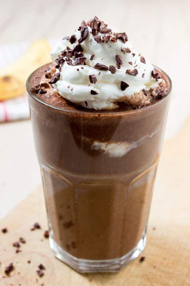Coffee breakfast smoothie in a tall glass topped with whipped cream and cacao nibs, a banana behind it