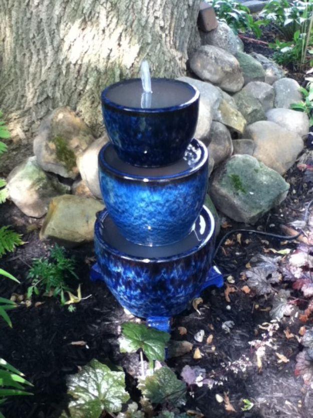 Three cobalt blue glazed pots stacked into a water fountain under a tree, ringed with mossy garden stones