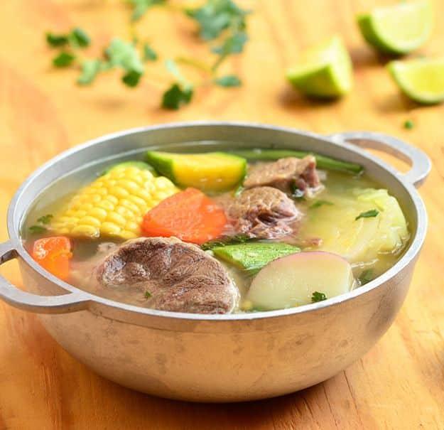 Caldo de res in a metal pot with beef shank, corn on the cob, carrots, potatoes, squash and lime wedges