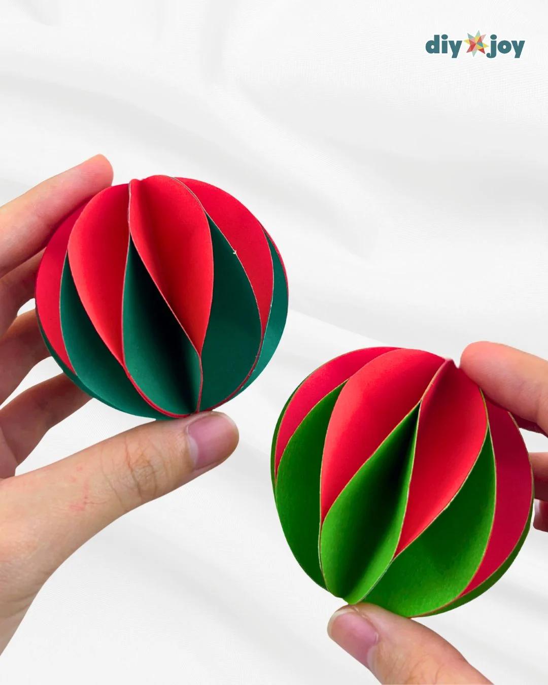 DIY Crafts - two hands holding up two finished paper ball ornaments in different red and green color combinations against a white backdrop