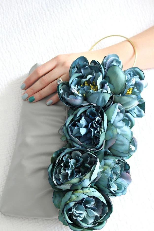 Grey vinyl clutch purse covered in dense turquoise faux peonies with a brass hoop handle, a hand resting beside it