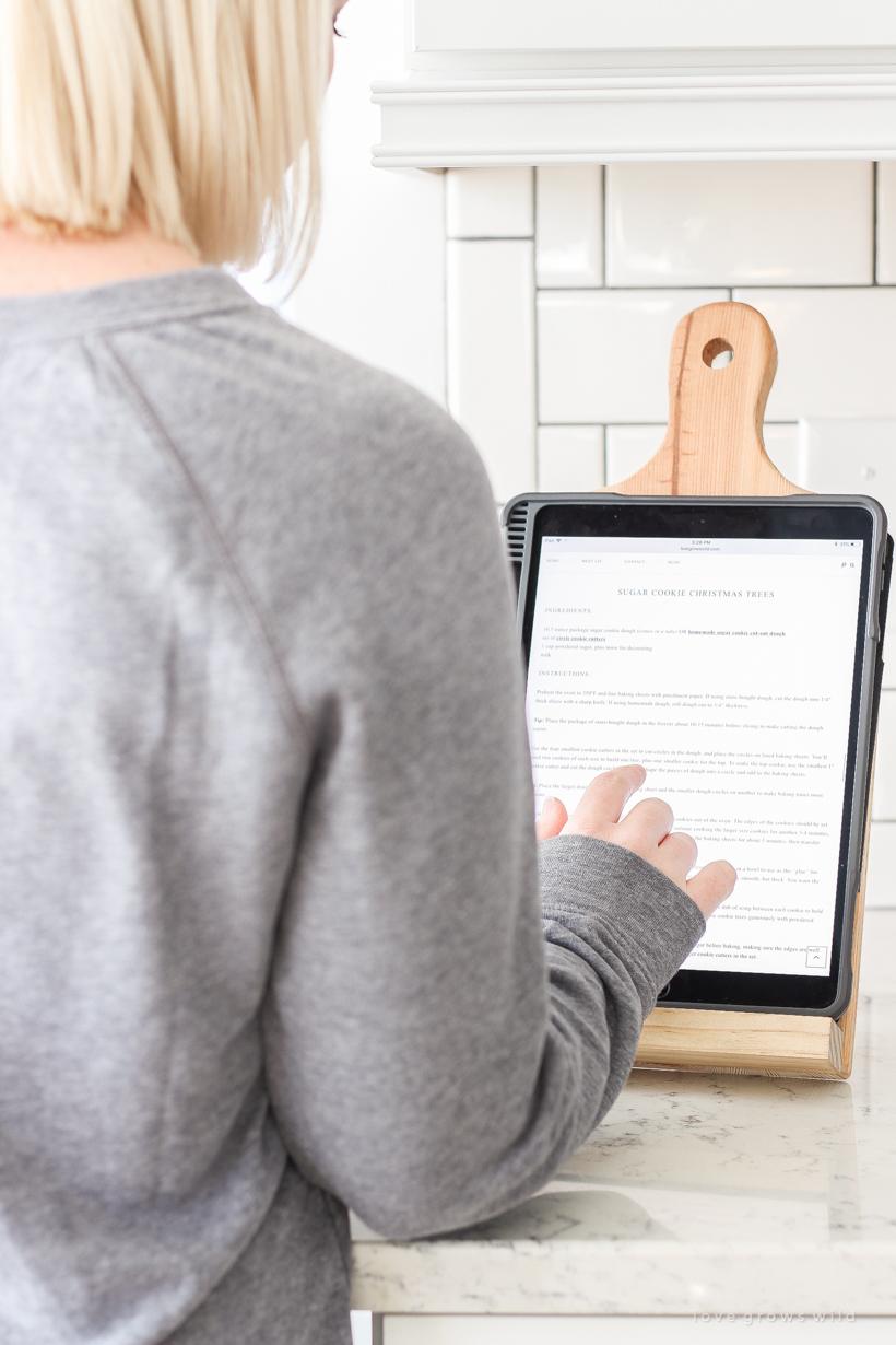 A wooden cutting board tablet holder on a white kitchen counter holding a tablet with a cookie recipe as a woman reads it