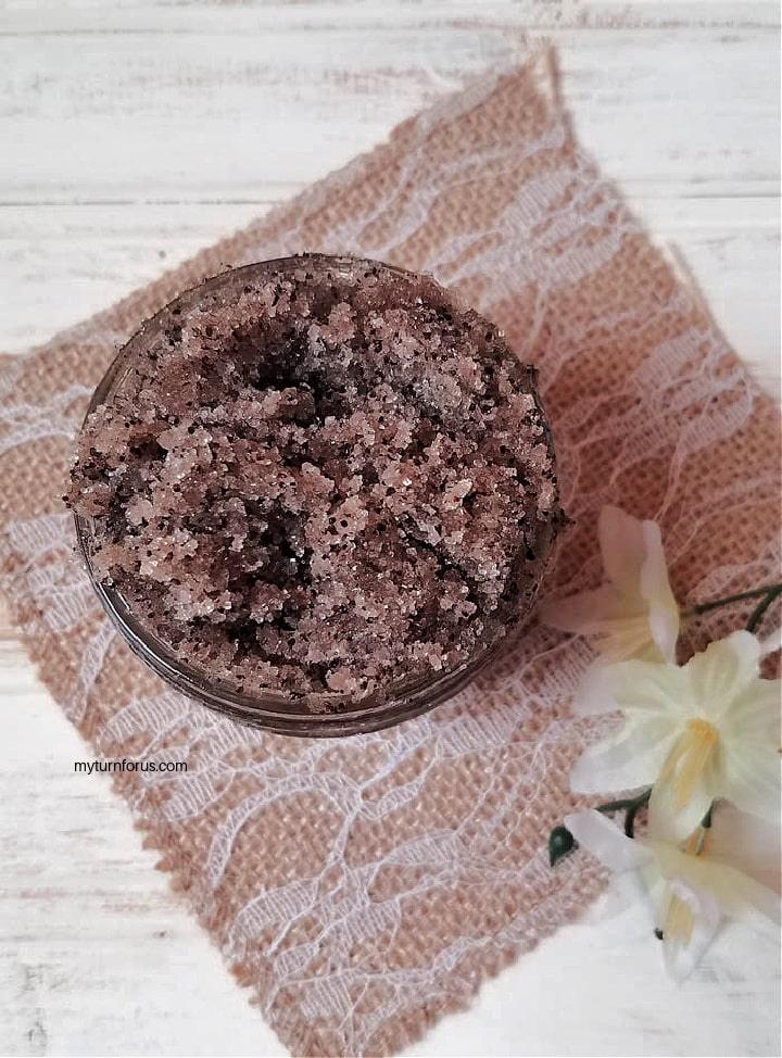 Small glass jar of brown coffee sugar scrub on a lace and burlap runner with a white flower beside it