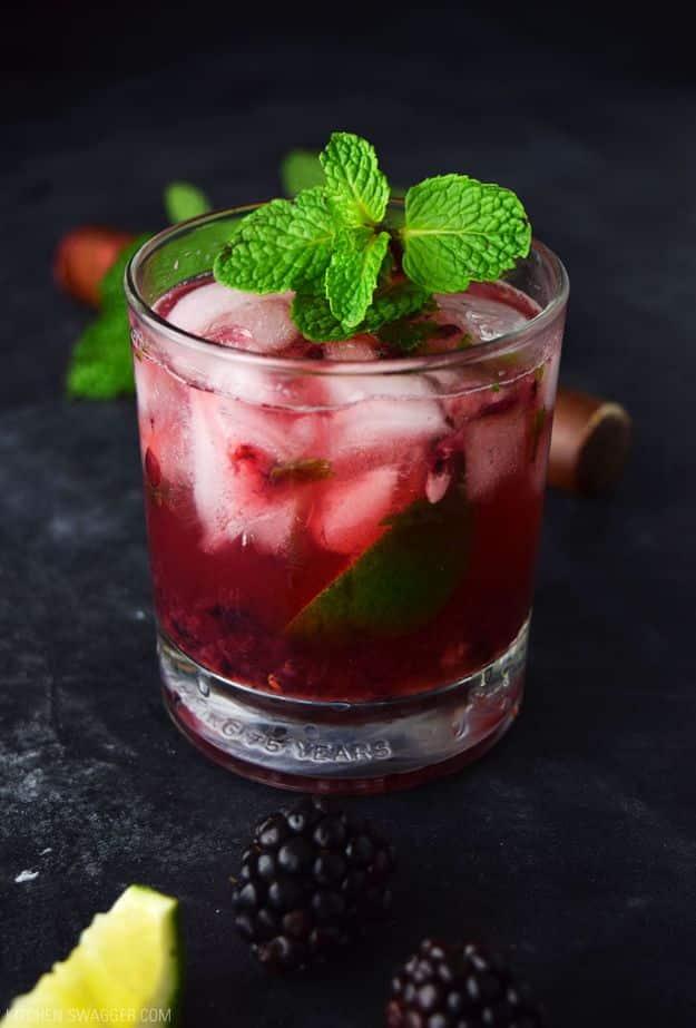 Short glass of blackberry bourbon smash over ice with mint, lime and fresh blackberries on a dark surface