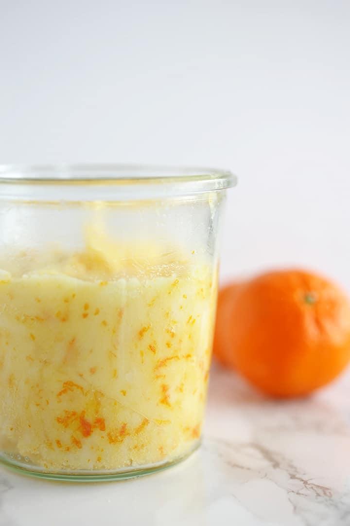 Glass jar of pale yellow sugar scrub flecked with orange zest, sitting on marble beside a whole mandarin